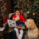 Woman reading a christmas story to a little girl in front of a Christmas tree with family dog.